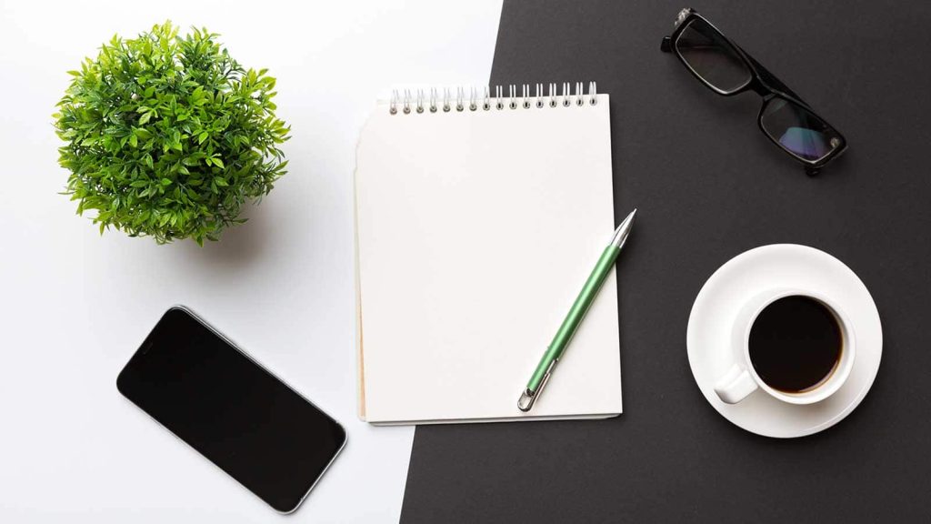 A modern office desk table with a notebook, smartphone, and other supplies and a cup of coffee. Blank notebook page for your design, top view flat lay.